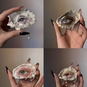 Op de afbeelding: Vier afbeeldingen van oesterschelp kunstwerken, elk met een gedetailleerd oog erin. De ogen hebben verschillende kleuren en zijn versierd met parels en parelkettingen. De schelpen worden vastgehouden in handen met lange, zwarte nagels.
