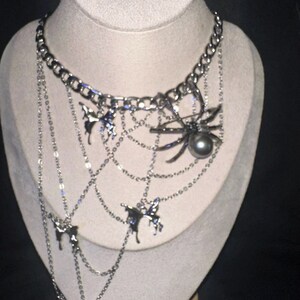 May include: Silver-toned necklace with a spider and web design. The necklace features a large spider pendant with a dark pearl, and several butterfly charms. The chain is made of multiple strands and hangs from a thick chain.