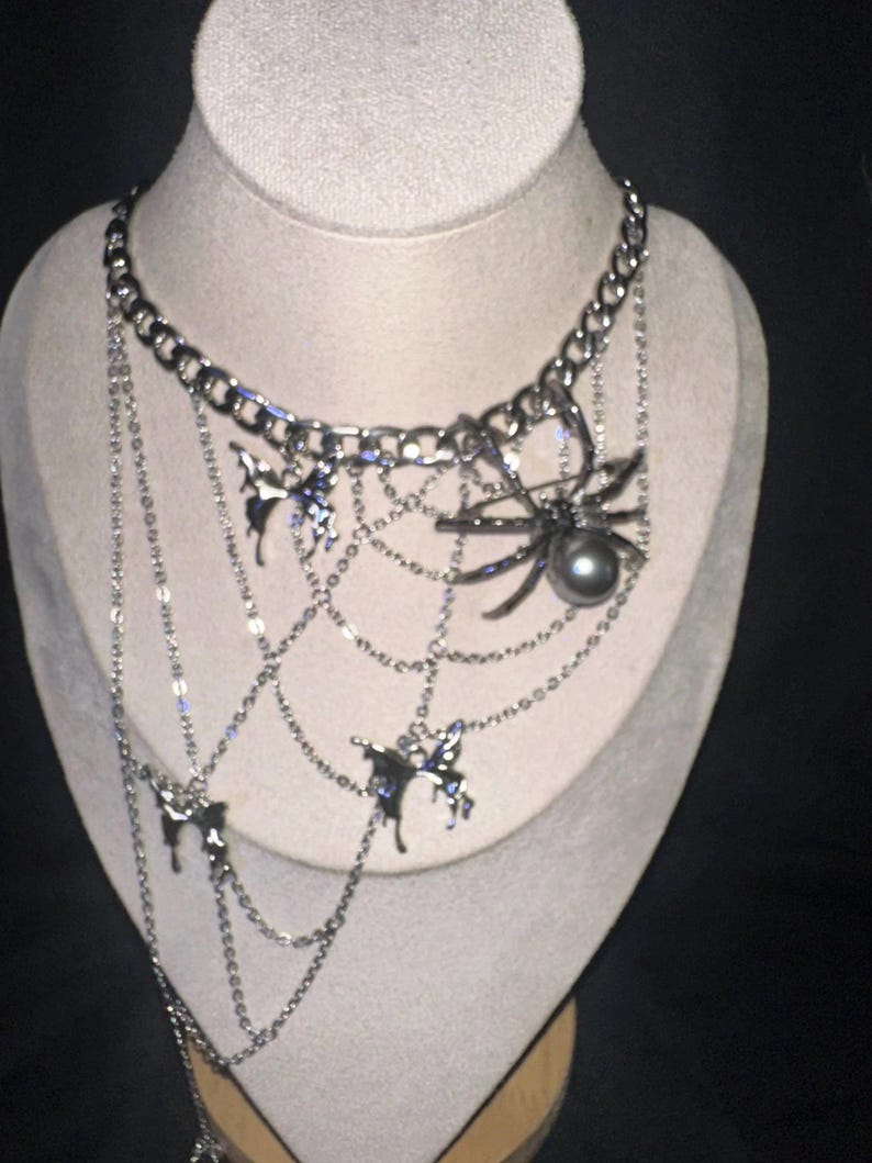 May include: Silver-toned necklace with a spider and web design. The necklace features a large spider pendant with a dark pearl, and several butterfly charms. The chain is made of multiple strands and hangs from a thick chain.