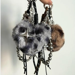 May include: Keyrings with fluffy, heart-shaped charms in grey and brown, decorated with silver-toned details such as crosses, stars, and beaded chains. The keyrings have black cords and silver key rings.