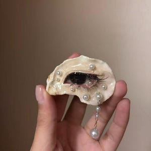 以下が含まれることがあります： 表面の中央にリアルな人間の目が描かれ、小さな白い真珠で囲まれた装飾用の牡蠣の殻。殻の下端からは、より大きな真珠が付いた銀色のチェーンが垂れ下がっており、暗い色のまつ毛がアクセントになっています。