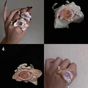 May include: A collection of five rings featuring realistic sculpted eyes set within irregular, shell-like structures. The designs incorporate small white pearls, textured metallic bands, and various iris colours including blue, brown, and iridescent purple tones.