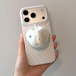 Puede incluir: Una funda de smartphone blanca con un patrón de puntos negros y un agarre tridimensional en forma de manzana blanca fijado en el centro. El agarre de manzana está decorado con pequeños cristales transparentes en forma de estrella y óvalo.