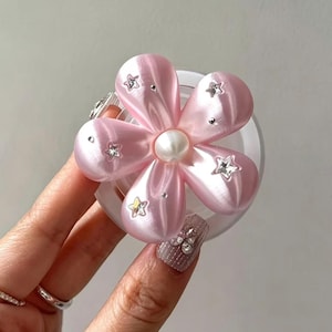 Pode incluir: Um suporte de telefone em forma de flor rosa com cinco pétalas lisas e arredondadas e um detalhe central de pérola branca. As pétalas são decoradas com pequenos pingentes de estrela prateados incrustados e minúsculos pontos metálicos.