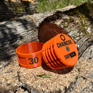 May include: Two orange 3D-printed objects. One is a cylindrical container with the number "30" printed on it. The other is a stack of orange discs with the text "CHAINCOIL" and "SHARP" printed on it. The objects are on a wood surface.