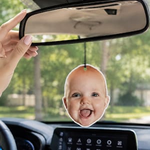 May include: A car air freshener hanging from a rearview mirror, featuring a die-cut, high-resolution photographic print of a smiling baby's face with light skin, blue eyes, and a small amount of hair, suspended by a thin black elastic cord.