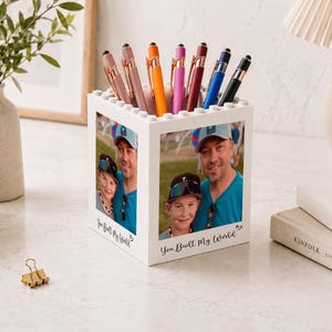 Puede incluir: Un portalápices cuadrado blanco con una textura de bloques de construcción en el borde superior. Los paneles frontal y lateral muestran una fotografía impresa de una persona y un niño. El texto "You Built My World" está impreso en negro.