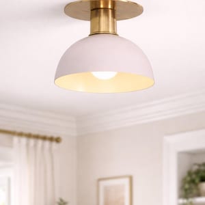 May include: A ceiling light fitting with a pale pink, domed shade and a brass-coloured base. The light bulb is visible inside the shade. The fitting is mounted on a white ceiling, with a glimpse of a window and a framed picture in the background.