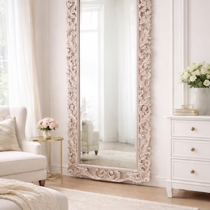 May include: A large, ornate, full-length mirror with a carved, cream-coloured frame. The mirror is rectangular with a decorative top and intricate floral and scrollwork detailing. The mirror is placed in a well-lit room with neutral tones.