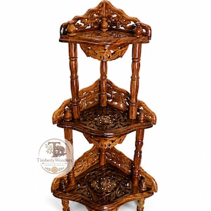 May include: A three-tiered brown wooden corner shelf featuring intricate floral carvings on each triangular surface and decorative turned pillars. The unit includes a circular logo on the left side with the text Timberly Woodora.