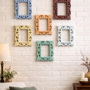 May include: Six ornate, rectangular picture frames in various pastel colors are mounted on a white brick wall. The frames are blue, peach, red-brown, yellow, green, and white. The frames have detailed, carved designs.