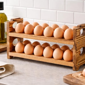 Könnte beinhalten: Ein hölzerner Eierhalter mit braunen Eiern. Der Halter hat zwei Ebenen und ist aus hellem Holz gefertigt. Eine Flasche Olivenöl und eine kleine Pflanze befinden sich im Hintergrund.