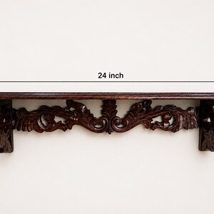 May include: A dark brown wooden wall shelf featuring intricate carved scrollwork and leaf patterns along the front support. The shelf measures 24 inches in width, 5 inches in height, and 5 inches in depth as indicated by the dimension labels.