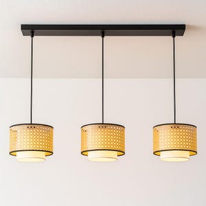 Puede incluir: Lámpara colgante de tres luces con pantallas cilíndricas. Las pantallas tienen un patrón tejido en color beige claro, con ribete negro y parte inferior blanca. La lámpara está suspendida de una base rectangular negra.