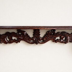 May include: A dark brown wooden wall shelf featuring an elongated rectangular top surface and a decorative apron underneath. The support brackets and the central section are intricately carved with detailed leaf and scroll patterns.
