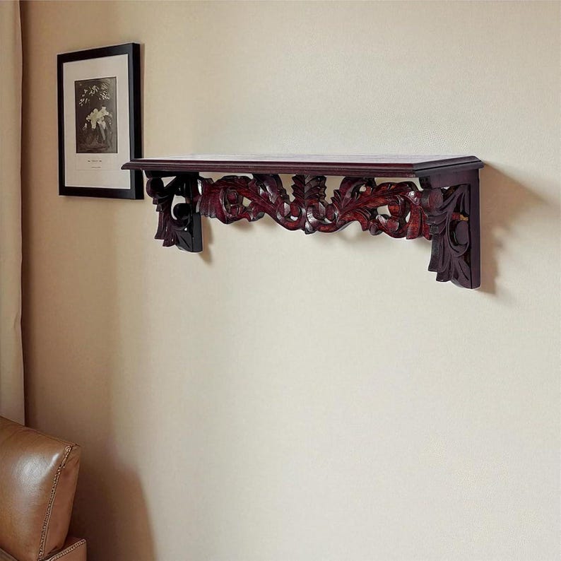May include: A dark brown wooden wall shelf featuring intricate, carved floral and scrollwork detailing along the front and side brackets. The shelf has a smooth, flat top surface and is mounted horizontally against a plain, light-colored wall.