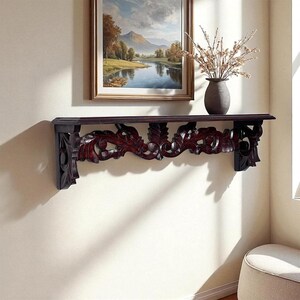 May include: A dark brown wooden wall shelf featuring intricate, carved floral and scrollwork patterns along the front apron. The shelf is mounted horizontally on a plain wall, providing a flat surface for display items.