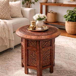 Puede incluir: Una mesa auxiliar redonda de madera marrón oscuro con intrincados diseños florales tallados. La mesa tiene una parte superior decorativa y una sección inferior con paneles tallados. Una bandeja dorada con un jarrón de flores blancas encima.