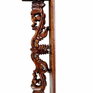 May include: A dark brown wooden wall shelf featuring an intricate vertical support bracket with carved leaf and scroll patterns. The shelf has a flat top and bottom surface with a straight back panel designed for mounting against a flat wall surface.