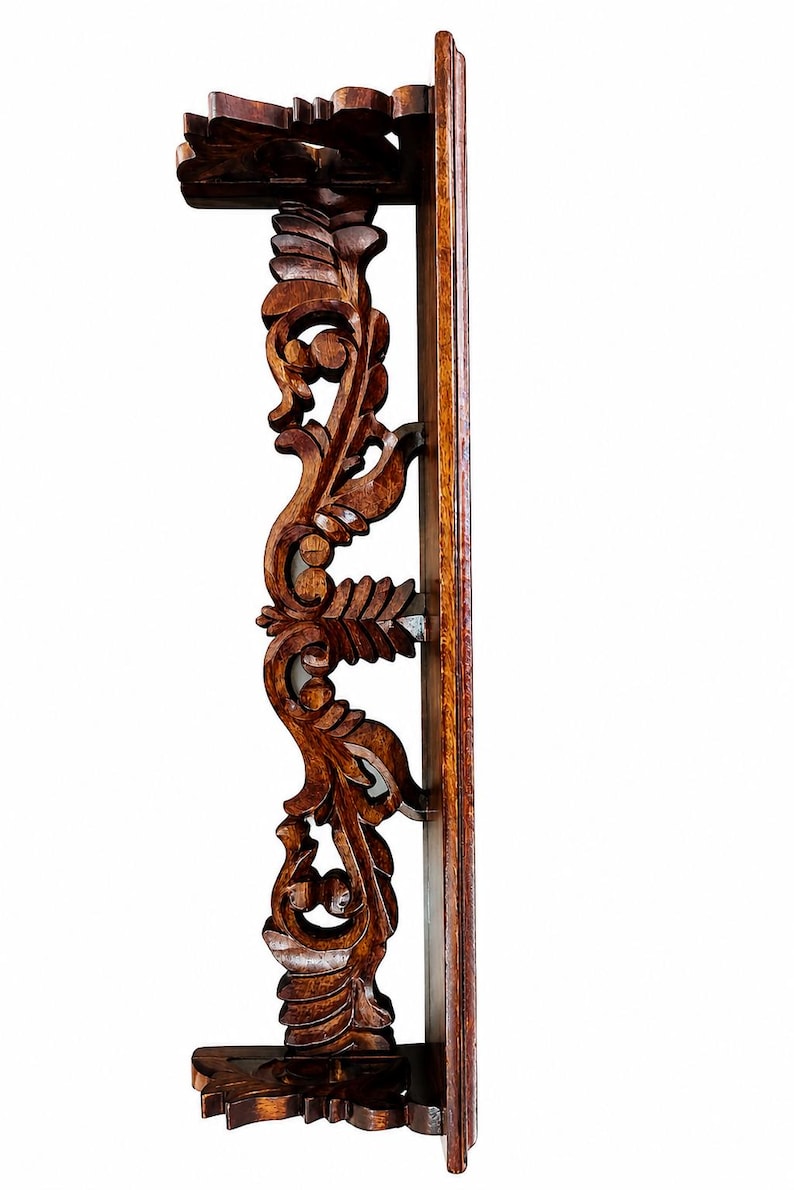 May include: A dark brown wooden wall shelf featuring an intricate vertical support bracket with carved leaf and scroll patterns. The shelf has a flat top and bottom surface with a straight back panel designed for mounting against a flat wall surface.