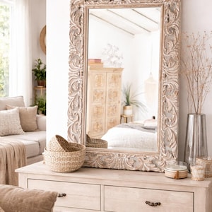 Peut inclure: Grand miroir rectangulaire avec un cadre en bois sculpté orné. Le cadre a une finition blanc cassé vieillie. Le miroir est posé sur une commode en bois clair, avec deux paniers tressés et des objets décoratifs.