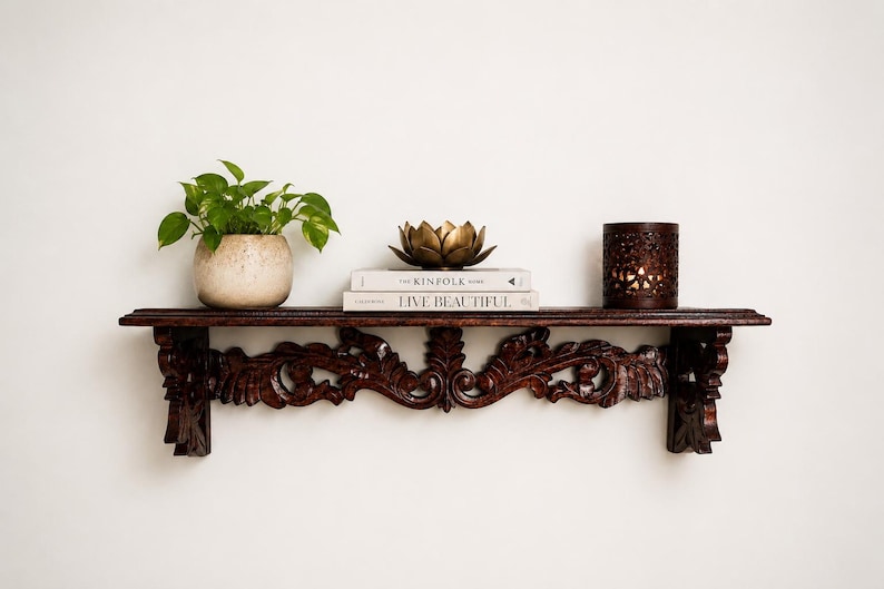May include: A dark brown wooden wall shelf with intricate carved scrollwork brackets. On the shelf sits a green plant in a speckled beige pot, two stacked books titled The Kinfolk and Live Beautiful, a gold lotus bowl, and a dark brown candle holder.