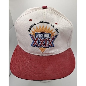 Puede incluir: Gorra de béisbol blanca y granate con el texto "SUPER BOWL XXIX" bordado en la parte delantera. La gorra tiene una visera granate y un botón en la parte superior. El texto "SUNDAY, JANUARY 29, 1995" también está bordado en la gorra.