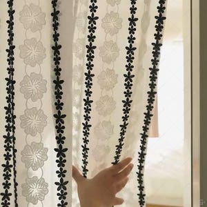 Puede incluir: Cortina blanca transparente con un estampado floral. La cortina presenta filas de bordados negros con forma de flor a lo largo de los bordes y entre los diseños florales. Una mano es parcialmente visible, sosteniendo la cortina abierta.