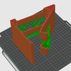 May include: A 3D-printed object in shades of brown, green, and red. The design features a rectangular structure with an angled top and internal supports. The object is on a dark gray surface with a grid pattern.