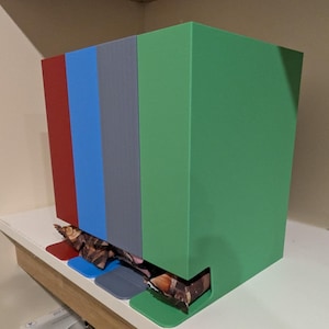May include: A multi-coloured rectangular box with red, blue, grey, and green panels. The box has a slot at the bottom, revealing a patterned item. The box is on a white surface.