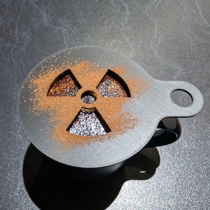 Könnte beinhalten: Eine schwarze Kaffeetasse mit einer grauen Schablone, die mit braunem Pulver bestäubt ist und ein Strahlungssymbol erzeugt. Die Schablone hat einen Griff mit einem kreisförmigen Loch. Der Hintergrund ist eine strukturierte graue Oberfläche.