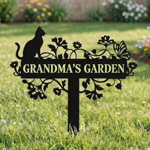 Puede incluir: Letrero de jardín de metal negro con el texto "Grandma's Garden". El letrero presenta una silueta de gato, flores y una mariposa. Está colocado en un jardín con hierba verde y flores coloridas.
