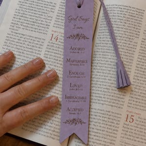 Puede incluir: Un marcapáginas rectangular de color morado claro con un borde inferior puntiagudo y una borla a juego en la parte superior. El marcapáginas presenta el texto God Says I am ADORED, MASTERPIECE, ENOUGH, LOVED, IRREPLACEABLE, ACCEPTED y motivos florales.
