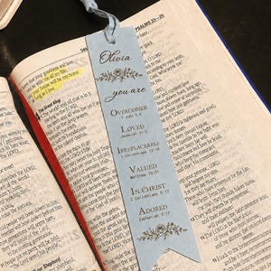Puede incluir: Un marcapáginas rectangular de color azul claro con una borla, que presenta el nombre Olivia y el texto you are OVERCOMER 1 John 5:4, LOVED Jeremiah 31:3, IRREPLACEABLE 1 Corinthians 12:18, VALUED Matthew 10:31, IN CHRIST 2 Corinthians 5:17, ADORED Zephaniah 3:17.