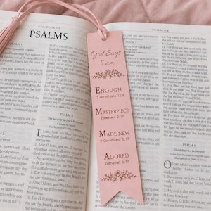 Puede incluir: Un marcapáginas rectangular de color rosa claro con una borla a juego, que presenta el texto God Says I am ENOUGH, MASTERPIECE, MADE NEW, ADORED y detalles florales, colocado dentro de un libro abierto con texto impreso en páginas blancas.