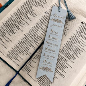 Puede incluir: Un marcapáginas rectangular de color azul claro con base puntiaguda y borla a juego. El marcapáginas presenta el nombre Hannah, adornos florales y el texto: you are HELD Isaiah 41:10, ADORED Zephaniah 3:17, NEVER ALONE Hebrews 13:5, NEEDED 1 Corinthians 12:21, ACCEPTED Romans 15:7, HEARD Jeremiah 33:3.