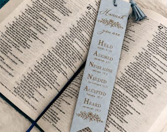May include: A light blue rectangular bookmark with a pointed bottom and a matching tassel. The bookmark features the name Hannah, floral accents, and the text: you are HELD Isaiah 41:10, ADORED Zephaniah 3:17, NEVER ALONE Hebrews 13:5, NEEDED 1 Corinthians 12:21, ACCEPTED Romans 15:7, HEARD Jeremiah 33:3.