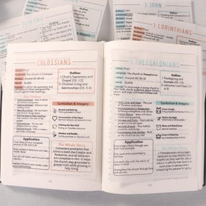 May include: Open book with pages displaying text from the Bible, including the books of Romans, Colossians, 1 Thessalonians, and 1 Corinthians. Several index cards with text are scattered around the book.