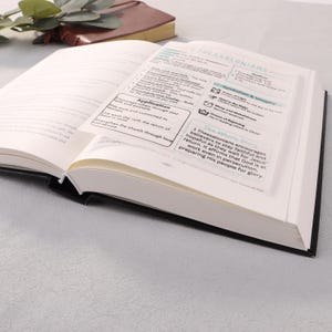 May include: An open book with black edges, featuring a clear overlay with text about Thessalonians. The overlay includes sections titled "Application" and "The Whole Story." A brown book and greenery are visible in the background.