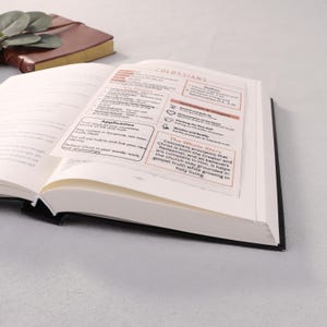 May include: An open book with black covers, displaying pages with text and highlighted sections. The text includes the word "Colossians" and other religious terms. A brown leather-bound notebook is visible in the background.