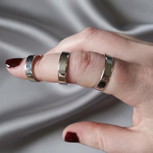 May include: Three silver rings are worn on a finger. The rings are simple bands with a polished finish. The nail on the fingertip is painted with dark red nail polish. The background is a soft, gray fabric.