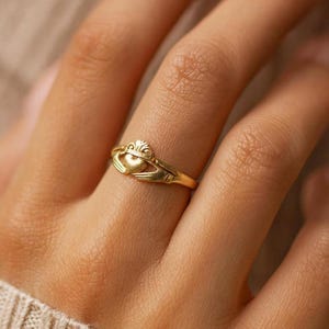 May include: A gold Claddagh ring, with a heart held by two hands and a crown. The ring is displayed on a finger, highlighting the detailed craftsmanship. The ring's design represents love, loyalty, and friendship. The ring is a classic piece of jewellery.