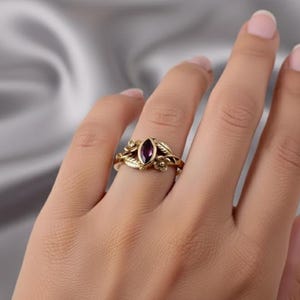 Könnte beinhalten: Ein goldfarbener Ring mit einem tiefvioletten, marquiseförmigen Edelstein. Der Ring hat ein Blumen- und Blätterdesign mit filigranen Details um den Edelstein und das Band. Der Ring befindet sich an einem Finger.