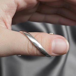 以下が含まれることがあります： カーブしたデザインのシルバーカラーの親指リング。リングは親指を包み込み、親指の上部と下部に小さな接続部分があります。リングは人の親指に表示されています。