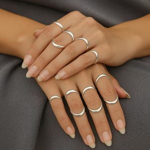 Puede incluir: Dos manos exhiben una colección de anillos plateados. Cada anillo tiene un diseño curvo. Los anillos son lisos y brillantes, reflejando la luz. Las manos se colocan sobre un fondo gris, resaltando las joyas.