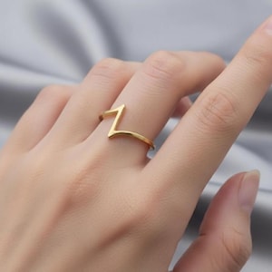 May include: A gold-coloured ring with a zigzag design. The ring is worn on a finger, and the design creates a modern, minimalist aesthetic. The ring's smooth surface reflects light, highlighting its metallic sheen. The ring is a piece of jewellery.