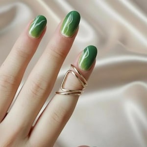 May include: A hand with green ombre nail polish and a gold geometric ring. The ring features a triangular design with two additional bands. The nails have a white swirl design.