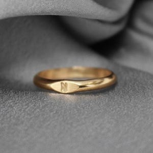 May include: A gold-colored ring with a rounded band and a flat, rectangular face. The face of the ring has the letter "N" engraved on it. The ring is set against a gray, textured background.