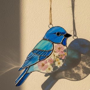 Puede incluir: Adorno de pájaro de vidriera en tonos azules y turquesas, con pico negro y diseño floral. El pájaro está suspendido por una cuerda. La sombra del adorno se proyecta sobre una pared de color crema.
