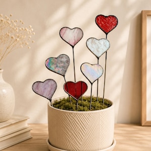 May include: A collection of heart-shaped glass garden stakes in various colors, including red, pink, and iridescent, are arranged in a white ceramic pot filled with green moss. The stakes have black metal stems and are displayed on a wooden surface.